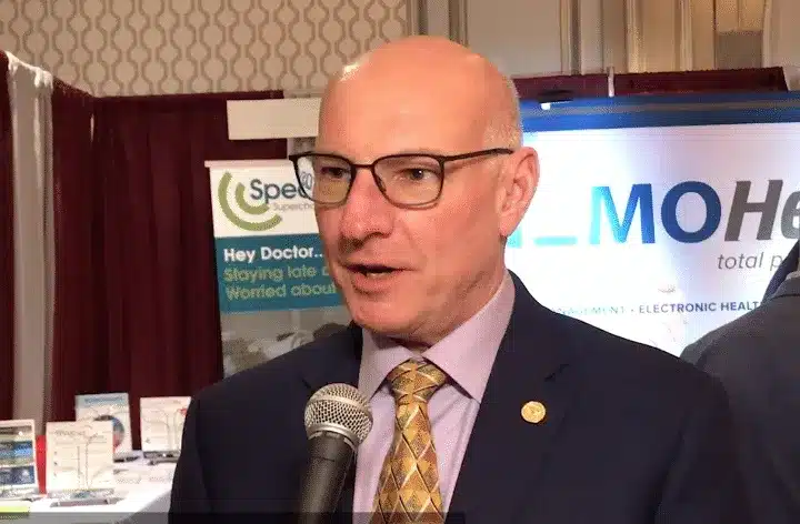 Bald man in glasses and a suit speaking into a microphone at a healthcare expo booth