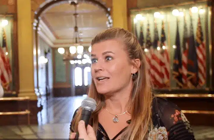 Woman speaking into a microphone in a historic hallway lined with flags and bright lights