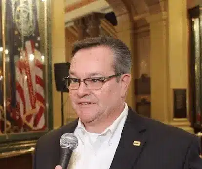 Man in glasses speaking into a microphone inside a government building with flags in the background